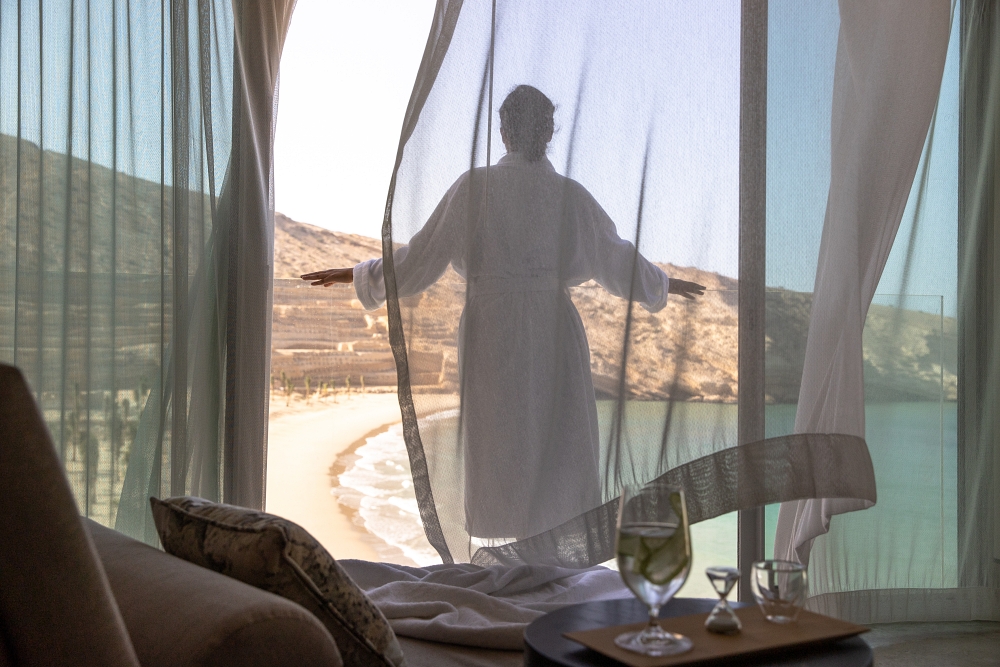 A woman standing in front of a window overlooking Muscat Bay with curtains billowing behind her