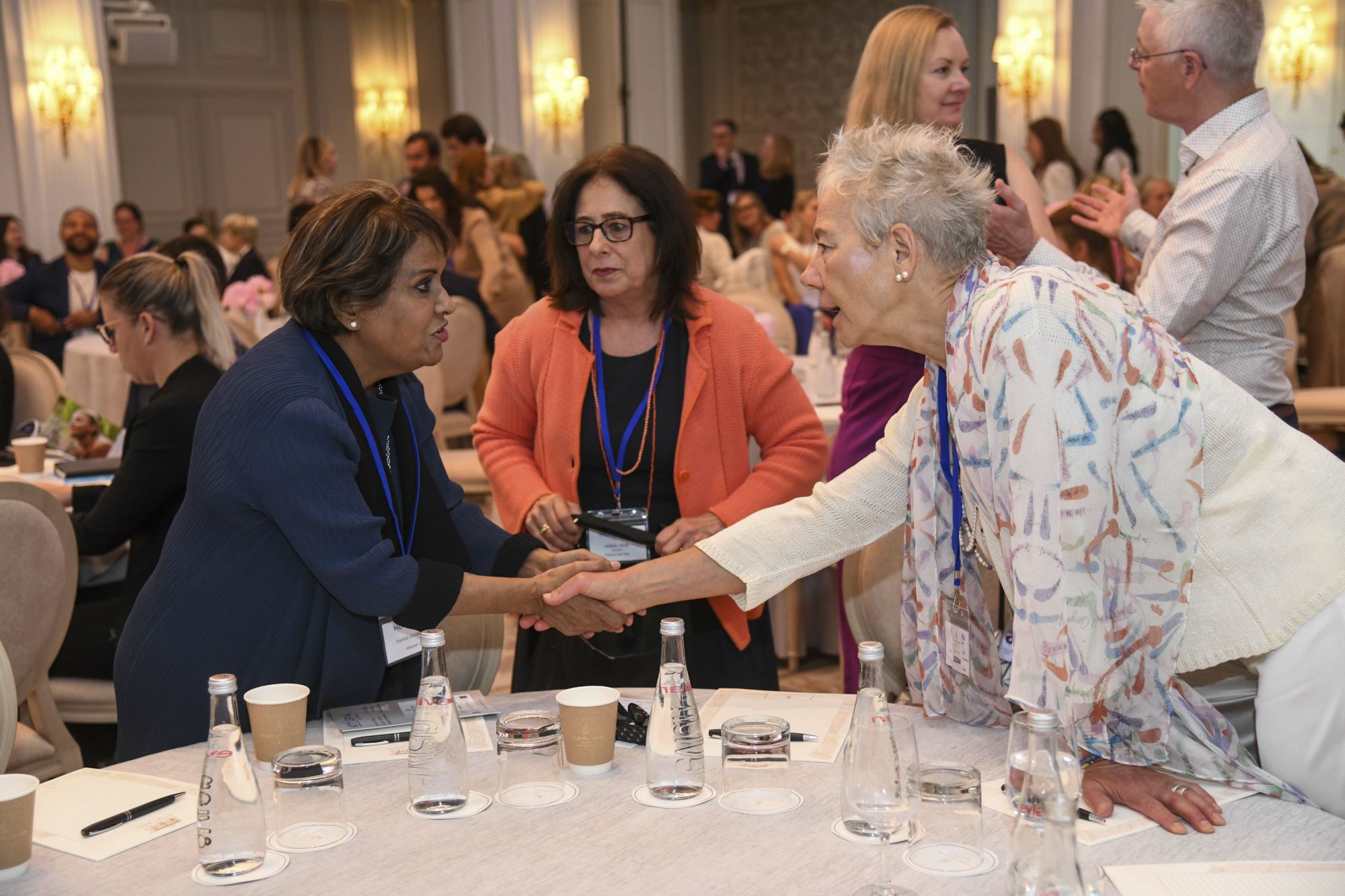 Three women greet each other across a table in a conference hall