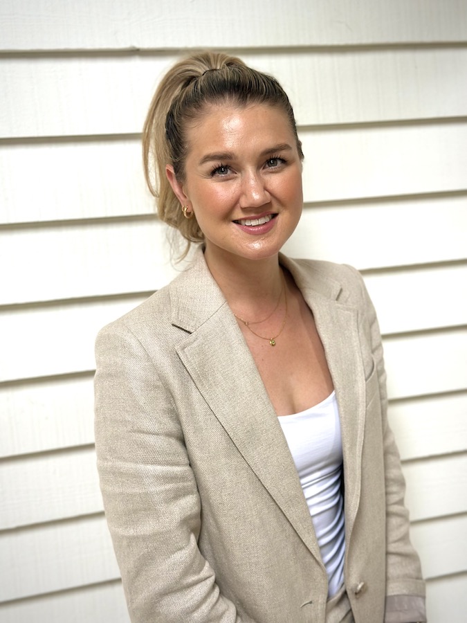 woman smiling at camera wearing a white top and beige jacket