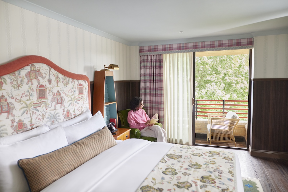 Woman sitting by the bed in a hotel room looking out of the window with a book in her lap